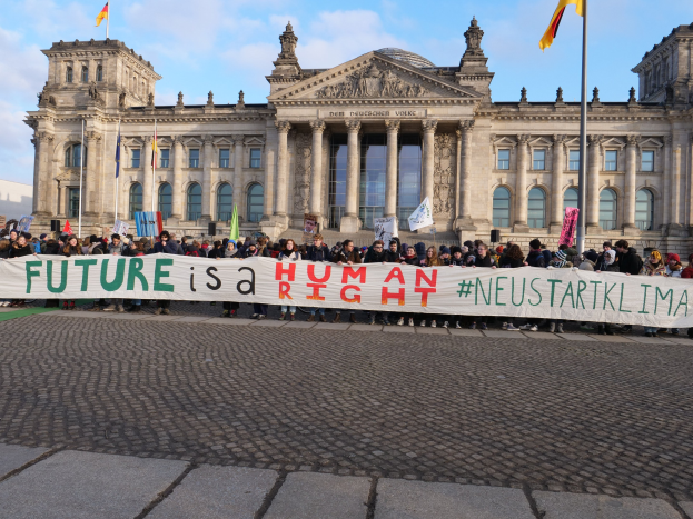 Gruppe von Menschen mit einem Banner "Zukunft ist ein Mensch" vor dem Reichstagsgebäude in Berlin, Deutschland, mit sichtbaren architektonischen Details und Flaggen im Hintergrund bei bewölktem Himmel.