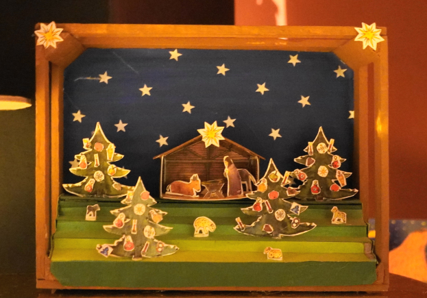 Eine hölzerne Krippe in einer Schachtel auf einem Tisch, mit einer Lampe links und einer Wand im Hintergrund, dekoriert mit Weihnachtsbäumen, Tieren und einem Haus.