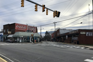 Stadtstraße mit Ampel an Kreuzung, Fahrzeuge auf der Straße, Gebäude mit Namensschildern, Strommasten mit Drähten, Bäume und bewölkter Himmel im Hintergrund.