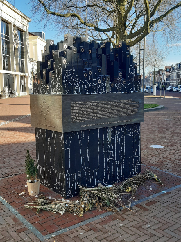 Denkmal in einem Stadtplatz gewidmet an Holocaust-Opfer, mit einem Baum davor, einem Topfgewächs, verstreuten trockenen Blättern, umgeben von Gebäuden, Fahrzeugen, Pfählen, Bäumen und Himmel.
