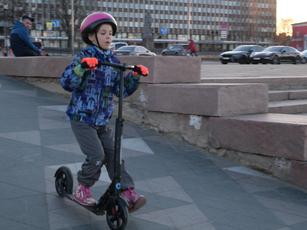Ein junger Junge fährt mit einem Helm und Handschuhen auf einem Gehweg mit einer E-Scooter, mit Stufen, Fahrzeugen, Menschen, Bäumen, Pfählen, Brettern, Gebäuden und einem klaren blauen Himmel im Hintergrund.