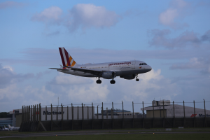 Germanwings Airbus A320-200 beim Start von Frankfurt Airport, mit Gras, einem Zaun, Gebäuden, Bäumen und einem bewölkten Himmel im Hintergrund.