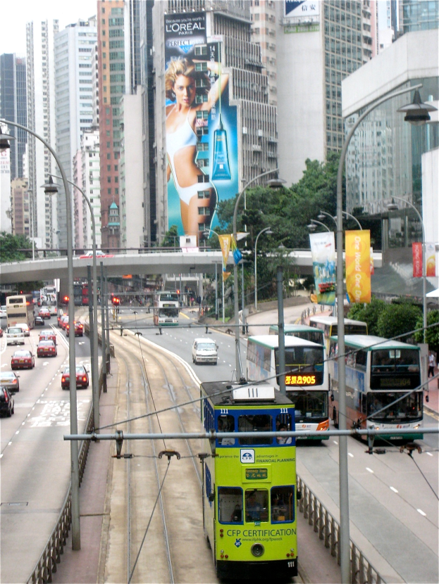 Eine belebte Stadtstraße mit verschiedenen Fahrzeugen, darunter Doppeldeckerbusse, Autos und andere Fahrzeuge, umgeben von hohen Gebäuden, Polen, Lampen, Schildern, Bannern, Bäumen und einer Brücke unter einem klaren blauen Himmel, mit Text am unteren Rand des Bildes.