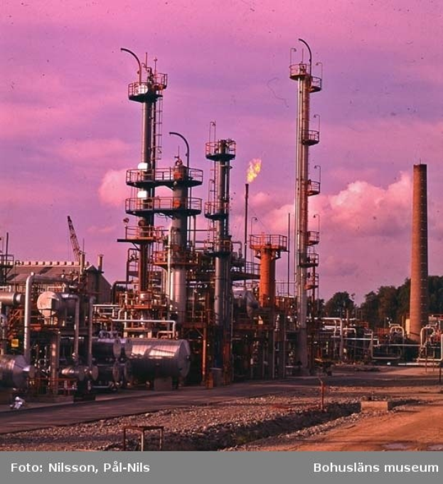 Industrielle Ölraffinerie mit ausgedehnten Rohrleitungen, Metallkonstruktionen, Containern, Bäumen, einem Gebäude, einem Kran und einem bewölkten Himmel, mit Text unten.