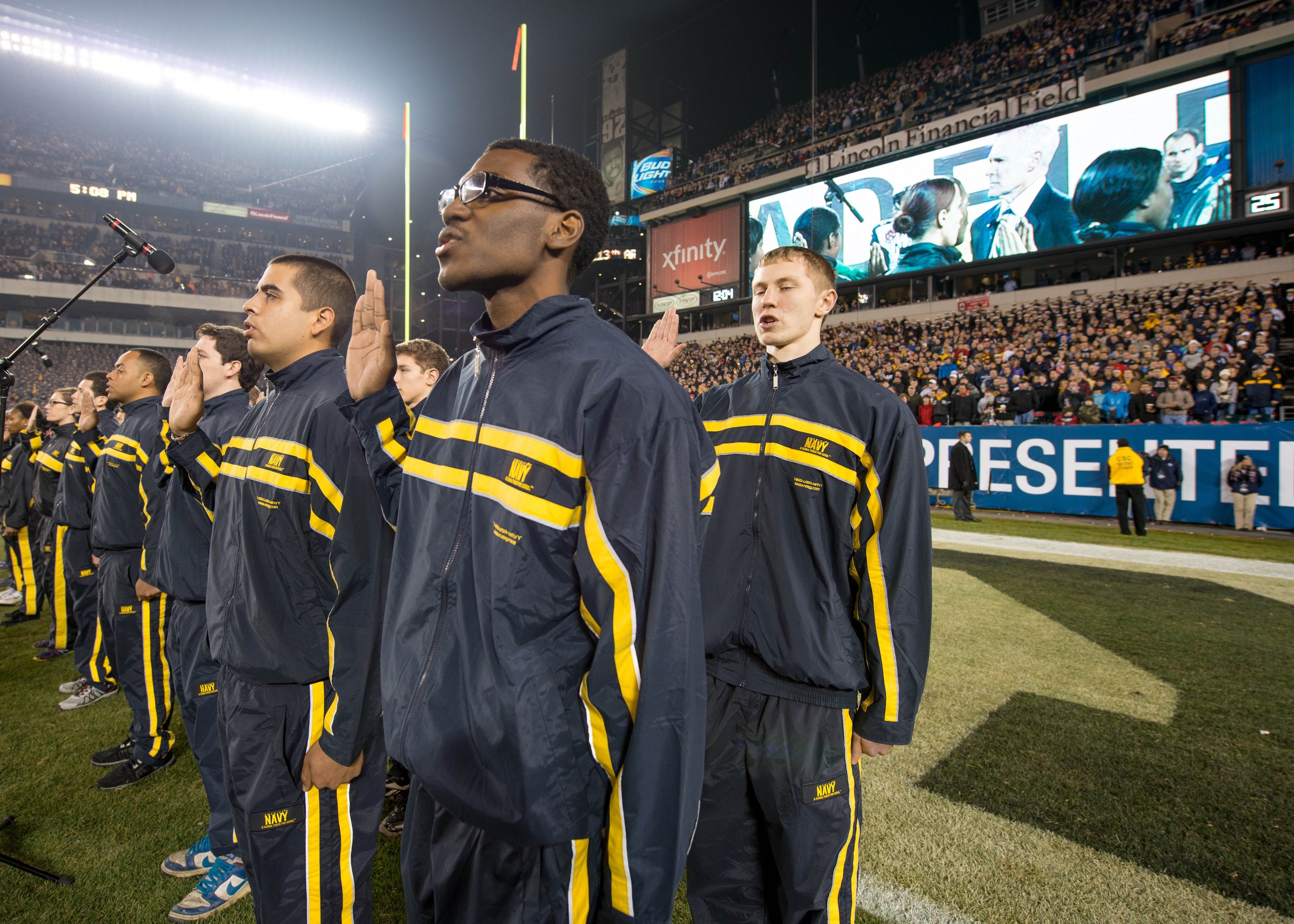 Eine Gruppe von Männern in blauen und gelben Jacken steht auf einem Fuballfeld, mit einer Stadionmenge, Schildern, Fahnen und Lichtern im Hintergrund und Mikrofonen, die eine Marschbandleistung anzeigen.
