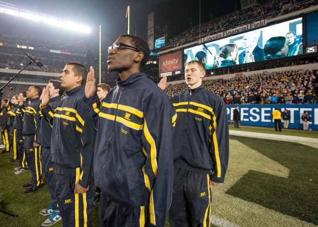 Eine Gruppe von Männern in blauen und gelben Jacken steht auf einem Fuballfeld, mit einer Stadionmenge, Schildern, Fahnen und Lichtern im Hintergrund und Mikrofonen, die eine Marschbandleistung anzeigen.