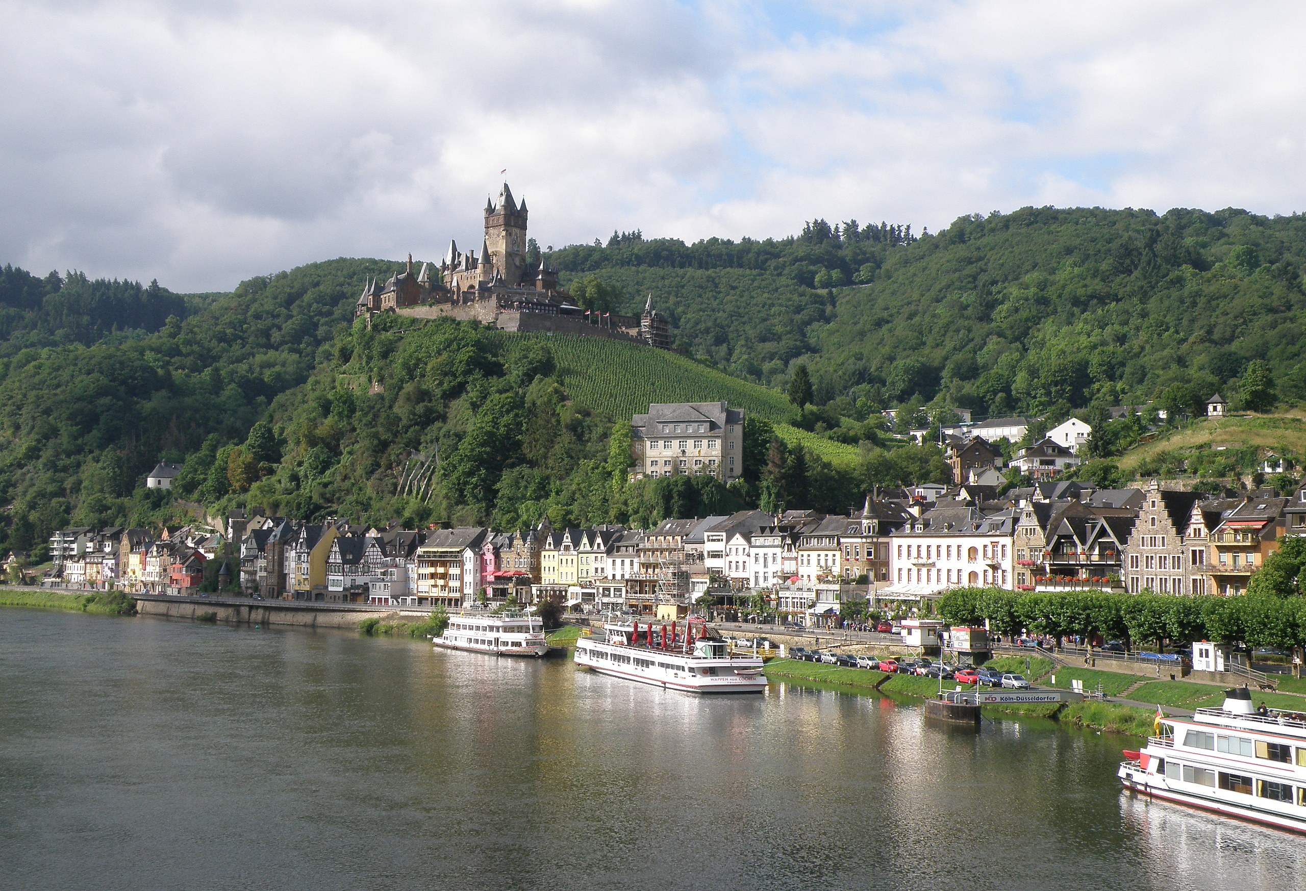 Ein malerischer Blick auf den Rhein in Deutschland mit einer Burg auf einem Hügel, Booten auf dem Fluss, Fahrzeugen auf einer näheren Straße und einer wolkigen Himmel.