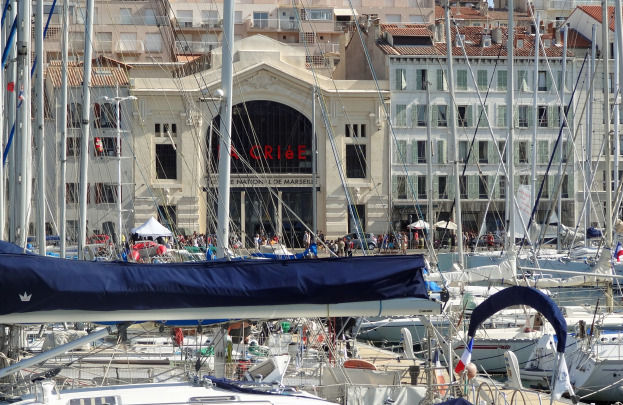 Ein Hafen mit zahlreichen Booten im Wasser, Gebäude mit Fenstern und einem Schild im Hintergrund sowie Zelte mit anwesenden Menschen.