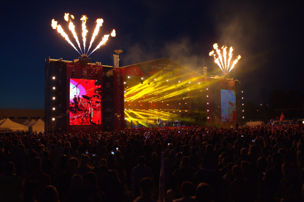 Großes Publikum bei einem Musikfestival mit beleuchteter Bühne, auf der Musiker spielen, umgeben von Bannern und Zelten links und Feuerwerk am dunklen Himmel.