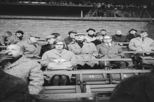 Eine Gruppe von Menschen, sowohl Männer als auch Frauen, sitzt in einem Stadion mit einer Wand und einigen stehenden Personen dahinter, dargestellt in Schwarz-Weiß.