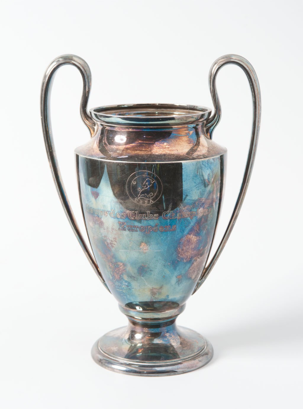 Der UEFA Champions League-Pokal, ein stahlerner Pokal mit zwei Handles und einem blauen und silbernen Farbschema, beschriftet mit "UEFA Champions League", gegen einen weißen Hintergrund dargestellt.
