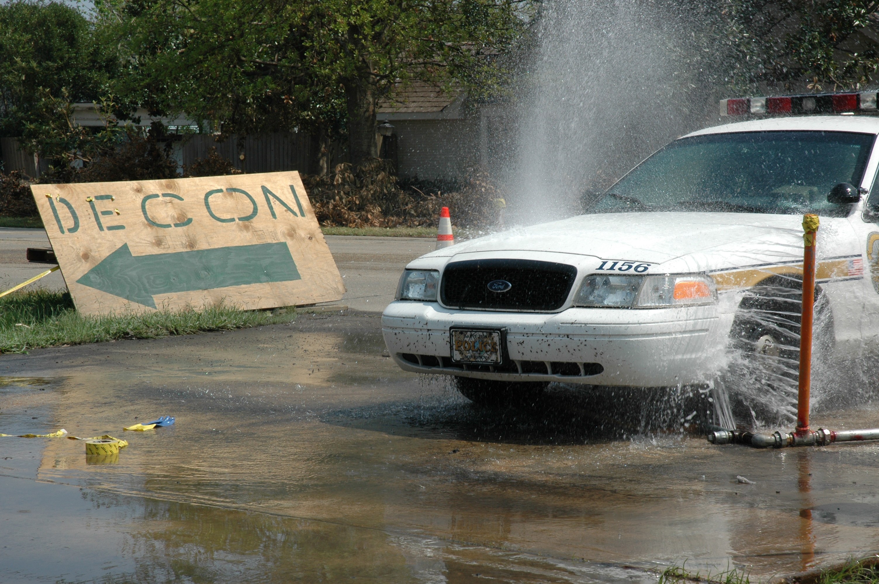 Ein Polizeiauto fährt durch eine Pfütze, mit einem Schild, einer Verkehrskegel und anderen Gegenständen auf dem Boden, vor einem Hintergrund aus Bäumen, Gebäuden und einem klaren Himmel.