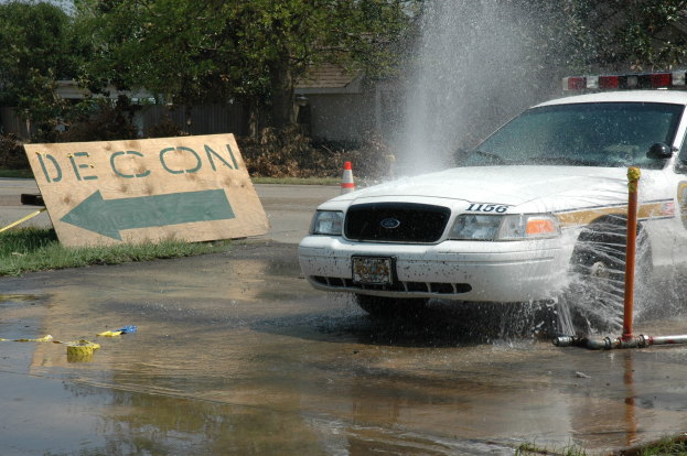 Ein Polizeiauto fährt durch eine Pfütze, mit einem Schild, einer Verkehrskegel und anderen Gegenständen auf dem Boden, vor einem Hintergrund aus Bäumen, Gebäuden und einem klaren Himmel.