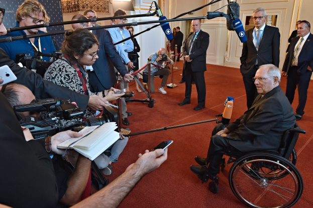 Ein Mann im Rollstuhl spricht mit Reportern, die Kameras, Mikrofone und Handys halten, vor einem Banner und Lichtern im Hintergrund.