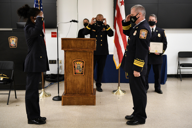 Gruppe uniformierter Personen in Masken vor einem Podium mit Mikrofon, umgeben von Fahnen und Möbeln, bei einer Vereidigung eines neuen Feuerwehrchefs, wobei eine Person ein Zertifikat hält.
