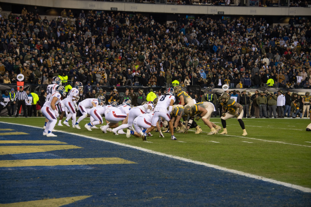 Ein Fußballspiel im Gange mit Spielern auf dem Boden in Sportkleidung und Helmen, von einem Stadionplatz aus mit Zuschauern, Geländern und Beschriftungen im Hintergrund.