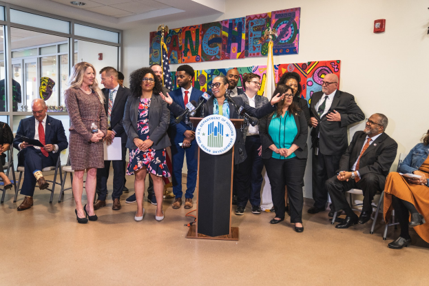 Gruppe von Menschen vor einem Rednerpult mit Mikrofon und einer Tafel mit dem Text "Sancho Stadtrat tritt dem Stadtrat bei", mit sitzenden und stehenden Teilnehmern, Flaggen, einem Feuerlöscher und Deckenleuchten im Hintergrund.