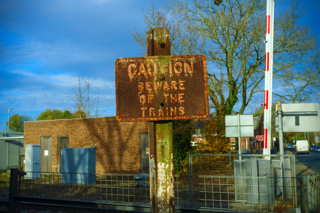 Vorsichtshinweis an einem Bahngeleise-Zaun mit Bäumen, Strommästen, einem Gebäude, Containern, Fahrzeugen auf einer Straße und einem bewölktem Himmel.