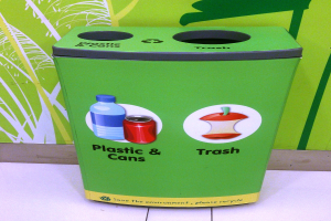 Ein grüner Müllcontainer voller Plastik und Dosen auf einem weißen Boden, mit einem Plakat an der Wand im Hintergrund.