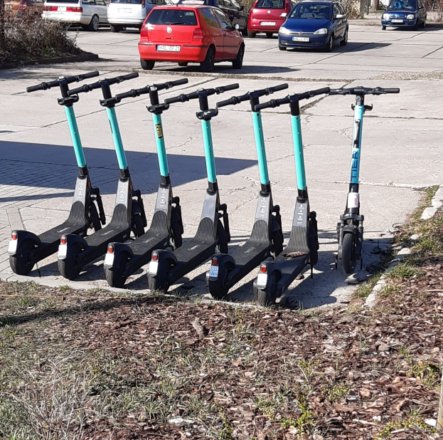 Eine Reihe von Elektro-Scootern, die auf einem grasbewachsenen Bereich neben einer Straße mit fahrenden Autos, Bäumen, Gebäuden und einem klaren blauen Himmel im Hintergrund geparkt sind.