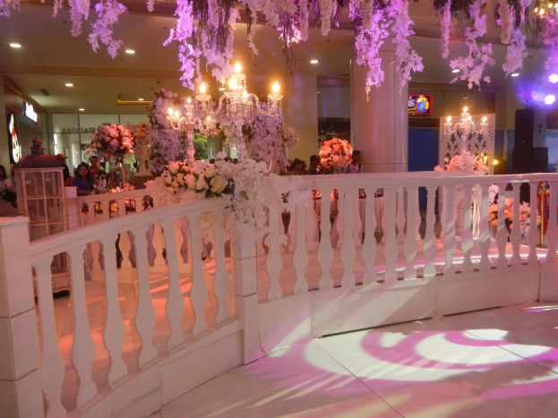 Weiße Reling mit floralen Dekorationen und hängenden Kronleuchtern bei einer Hochzeitsfeier, Menschen umringen die Reling, beleuchtete Säulen im Hintergrund und ein Bildschirm auf der rechten Seite.