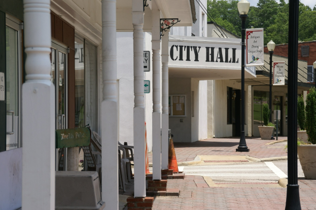 Ein Rathaus mit Gebäuden, Straßenlaternen, Straßenschildern, Stühlen, Topfpflanzen, Bäumen und einem bewölkten Himmel im Hintergrund.