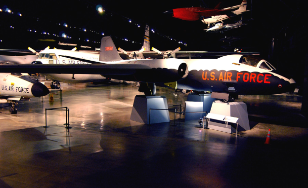 Gruppe von Kampfflugzeugen in einem Museum ausgestellt, beleuchtet von ├ťberkopfbeleuchtung gegen einen dunklen Hintergrund, mit St├Ąnden, Verkehrskegeln und anderen Gegenst├Ąnden auf dem Boden.