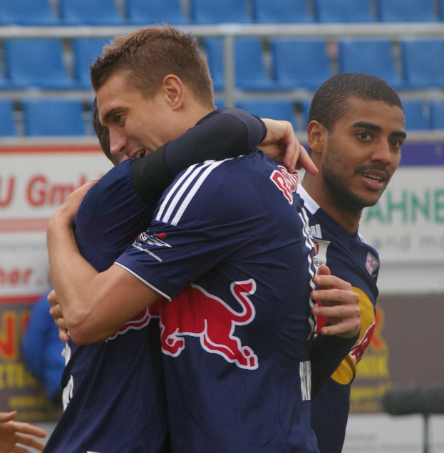 Zwei Fußballspieler umarmen sich auf einem Feld, mit einer Hand sichtbar auf der linken Seite und im Hintergrund einige Menschen, Schilder mit Wörtern und blaue Stühle.