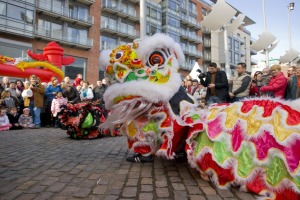 Ein lebendiges chinesisches Neujahrsfest in Amsterdam mit einem Löwen tanzen im Vordergrund und einer Menge Menschen, einige mit Kameras, die darum herum versammelt sind, und Gebäuden, Laternenmasten und einem klaren blauen Himmel im Hintergrund.