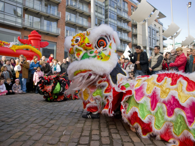Ein lebendiges chinesisches Neujahrsfest in Amsterdam mit einem Löwen tanzen im Vordergrund und einer Menge Menschen, einige mit Kameras, die darum herum versammelt sind, und Gebäuden, Laternenmasten und einem klaren blauen Himmel im Hintergrund.