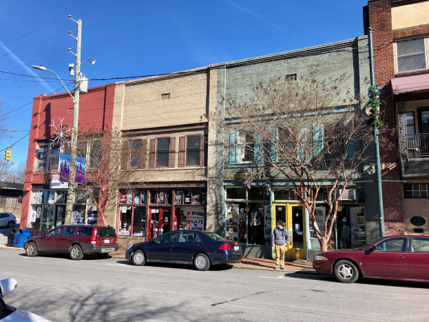 Eine Straße in einer Stadt mit geparkten Autos, Gebäuden, Bäumen, Laternen, Strommästen mit Käblein und einer Person, die auf dem Gehweg spaziert, unter einem klaren Himmel, darstellend eine gewerbliche Immobilie zum Verkauf zu $1,250,000.