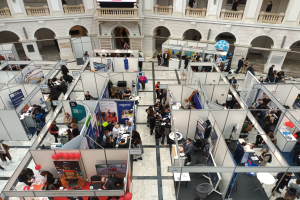 Eine große Gruppe von Menschen versammelt sich in einem Raum mit zahlreichen Ständen, Bannern, Tischen, Stühlen und Ballons, vor einem Gebäude mit Bögen, Säulen, Geländern und einem Bildschirm im Hintergrund.