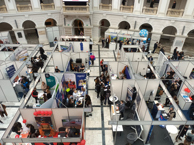 Eine große Gruppe von Menschen versammelt sich in einem Raum mit zahlreichen Ständen, Bannern, Tischen, Stühlen und Ballons, vor einem Gebäude mit Bögen, Säulen, Geländern und einem Bildschirm im Hintergrund.