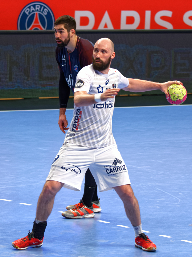 Zwei Männer spielen Handball auf einem Court, einer hält einen Ball, mit einer Tafel im Hintergrund, die 'Paris Saint-Germain vs Paris Saint Germain' anzeigt.