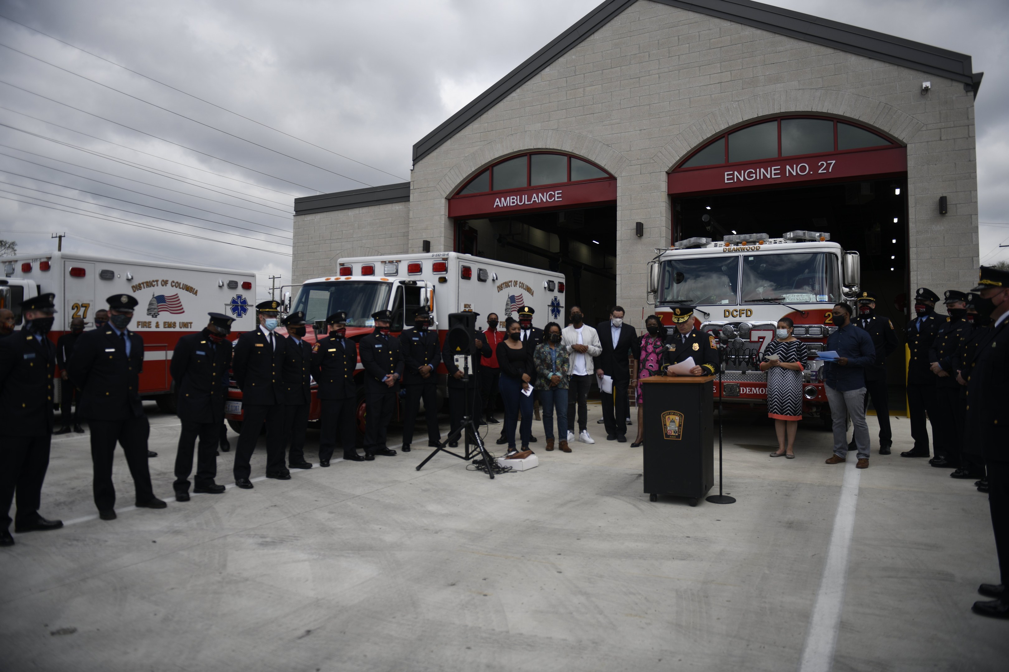 Gruppe von Menschen vor einer Feuerwache mit Feuerwehrautos, einem Podium, Mikrofon, Bäumen, Strommasten und einem klaren blauen Himmel im Hintergrund.