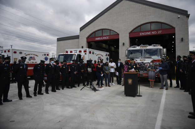 Gruppe von Menschen vor einer Feuerwache mit Feuerwehrautos, einem Podium, Mikrofon, Bäumen, Strommasten und einem klaren blauen Himmel im Hintergrund.