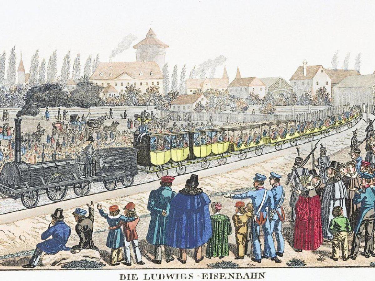 Eine Gruppe von Menschen, die um einen Zug auf Schienen mit Gebäuden und Bäumen im Hintergrund stehen und sitzen, beschriftet mit "Ludwigs-Eisenbahn, die erste Bahnstation in Deutschland."