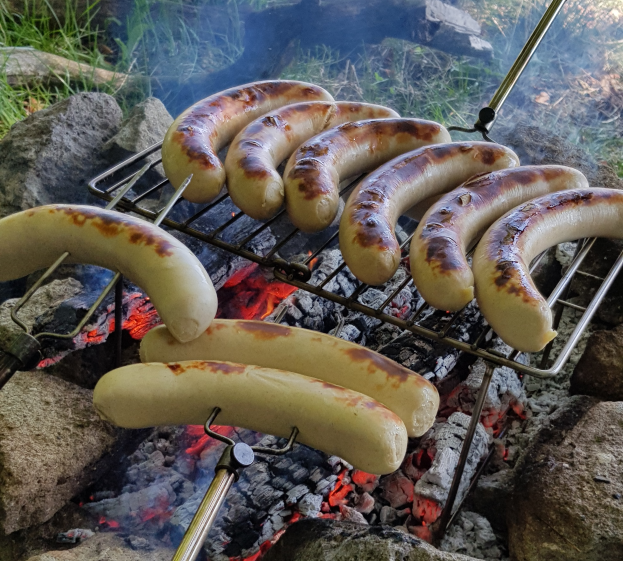 Würstchen auf einem Grill über einem Lagerfeuer, umgeben von Steinen und Gras.