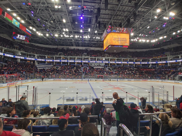 Eine große Menschenmenge in einer Hockeyarena, einige sitzen und einige stehen, schauen ein Spiel unter hellen Lichtern mit Bannern an der Decke.