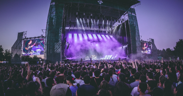 Eine gro├če Menge auf einem Musikfestival schaut auf einer beleuchteten Bühne mit Instrumenten und Bildschirmen, die vor einem Geb├Ąude, B├Ąumen und einem blauen Himmel stehen.
