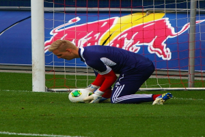 Ein Fußballspieler kniet auf dem Boden mit einem Ball vor einem Tor, umgeben von Gras und einem Netz, mit einem Banner mit Text und einem Bild einer Person im Hintergrund.