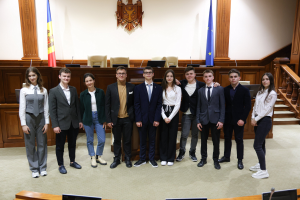 Gruppe von Menschen, die vor einem Gerichtssaal mit Monitoren und Mikrofonen auf Tischen und einer hölzernen Plattform mit Stühlen, Flaggen und einem Wandobjekt im Hintergrund posieren.