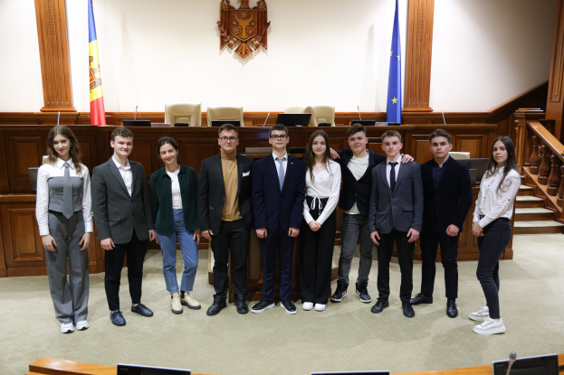 Gruppe von Menschen, die vor einem Gerichtssaal mit Monitoren und Mikrofonen auf Tischen und einer hölzernen Plattform mit Stühlen, Flaggen und einem Wandobjekt im Hintergrund posieren.