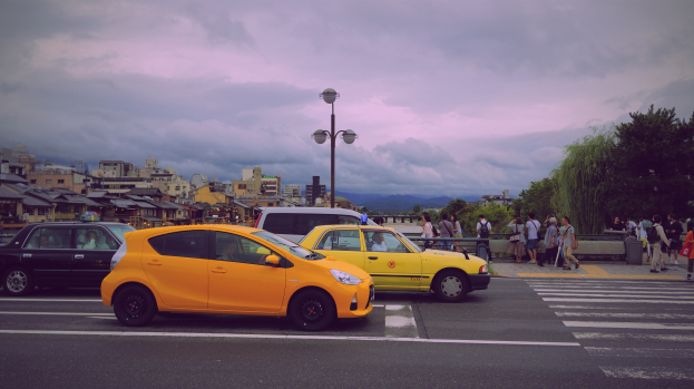 Zwei gelbe Taxis stehen auf der Seite einer Stadtstraße mit Passanten, Laternenpfählen, Bäumen, Gebäuden und einem bewölkten Himmel im Hintergrund.