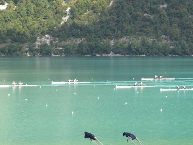 Gruppe von Menschen bei einem Ruderwettbewerb auf einem Gewässer, umgeben von Bäumen und einem Hügel im Hintergrund, mit über Stühle drapierten Kleidungsstücken unten.