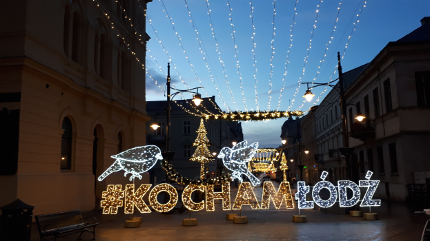 Eine belebte Stadtstraße in Krakau, Polen, dekoriert mit festlichen Weihnachtslichtern, gesäumt von Gebäuden, Straßenlaternen, Lichtern, Bänken und beleuchteten Dekorationen unter einem sichtbaren Himmel.