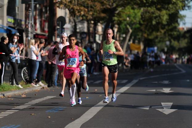 Gruppe von Menschen, die bei einem Marathon auf einer städtischen Straße laufen, mit Zuschauern auf der linken Seite, unscharfer Hintergrund mit Bäumen, Gebäuden, Pfählen, Schildern und einem Fahrrad.