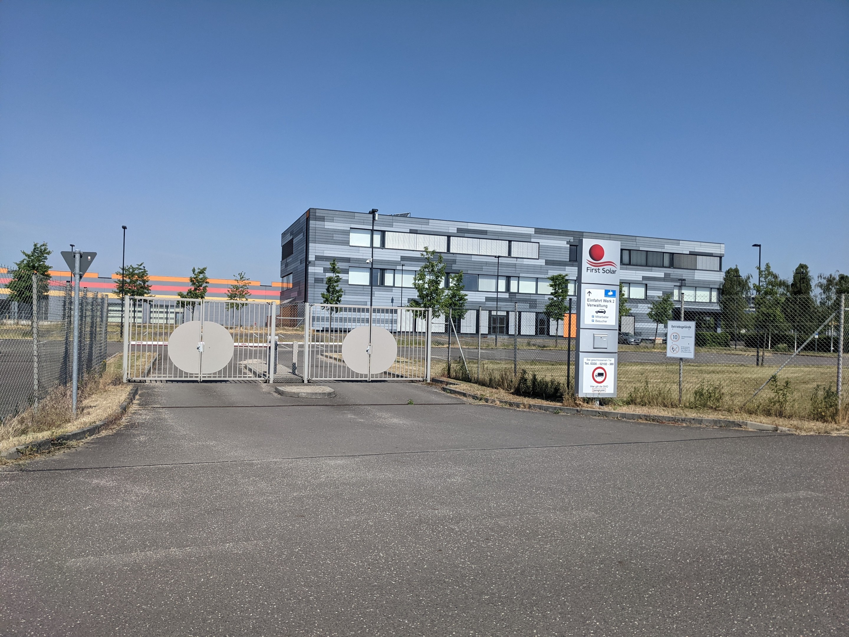 Großes industrielles Gebäude mit der Bezeichnung "First Solar" mit vielen Fenstern, umgeben von einem Metallzaun, umgeben von Bäumen, Pflanzen, Gras, Schildern, Straßenlaternen und einem Weg unter einem bewölkten Himmel.