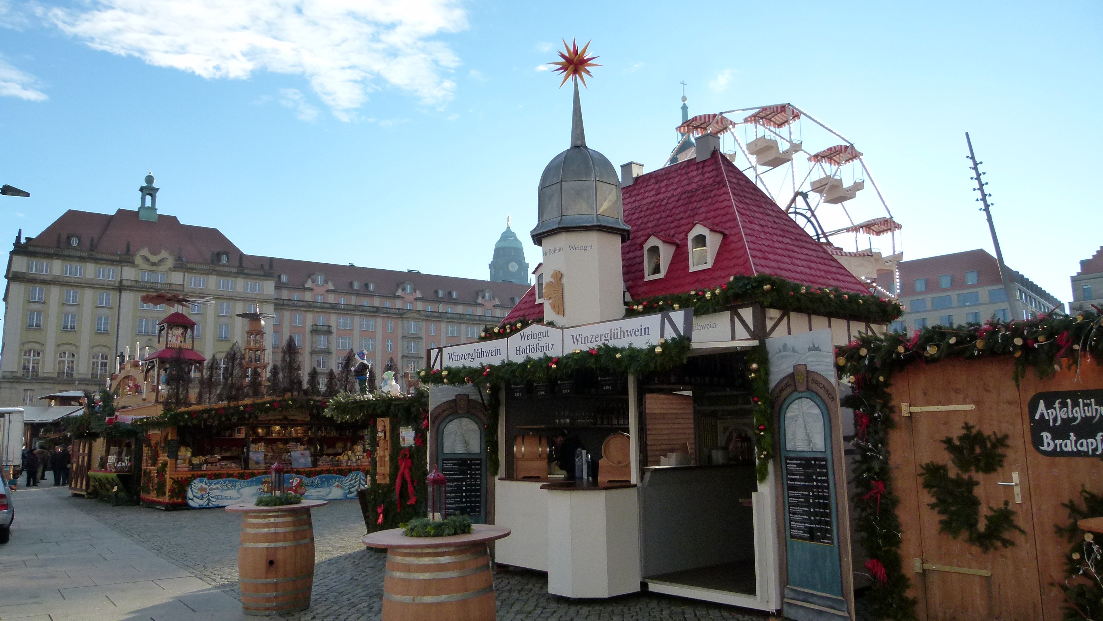 Ein geschäftiger Weihnachtsmarkt auf einem Stadtplatz mit geschmückten Bäumen, Gebäuden, Fahrzeugen, Passanten und bewölktem Himmel.
