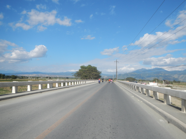 Eine Autobahn mit Motorradfahrern, umgeben von Geländern, Strommästen mit Drähten, Bäumen, Hügeln und einem bewölkten Himmel.